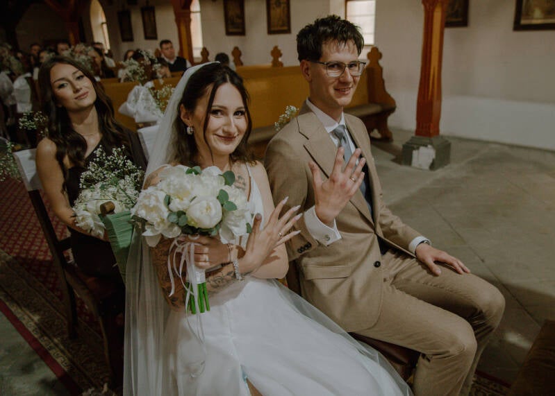 Fotografia ślubna Grudziądz – radosna para młoda pokazuje złote obrączki ślubne w kościele w Piaskach. Wiktoria z bukietem białych piwonii i Dawid w brązowym garniturze siedzą przed ołtarzem, uśmiechając się do aparatu w towarzystwie świadkowej.