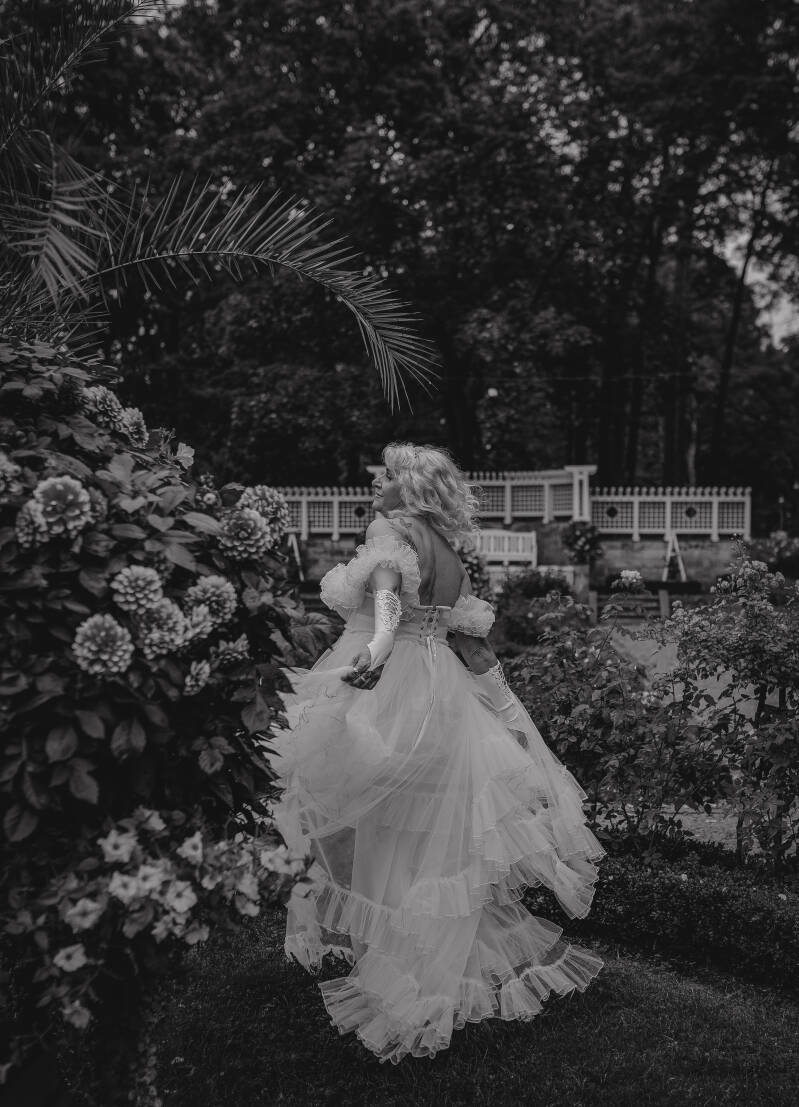 Plenerowa sesja urodzinowa dla kobiet kujawsko-pomorskie. Monochromatyczna fotografia sensualna i glamour w plenerze. Kobieta w długiej, tiulowej kreacji z autorskiej garderoby fotografki, pozująca dynamicznie tyłem wśród bujnych krzewów klombu kwiatowego
