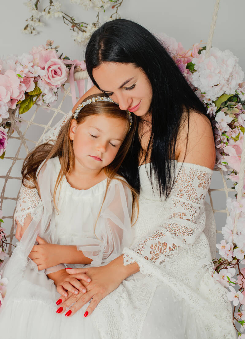 Mama w białej, koronkowej sukni boho z odkrytymi ramionami przytula córkę na huśtawce ozdobionej kwiatami podczas sesji w Grudziądzu