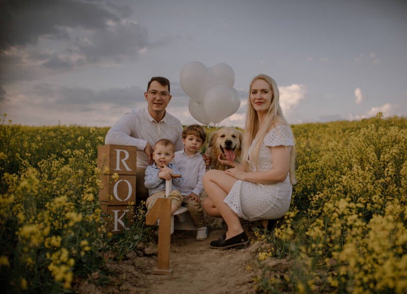 Rodzina pozuje w rzepaku z psem golden retrieverem, białymi balonami i drewnianymi dekoracjami na roczek – sesja urodzinowa w okolicach Grudziądza