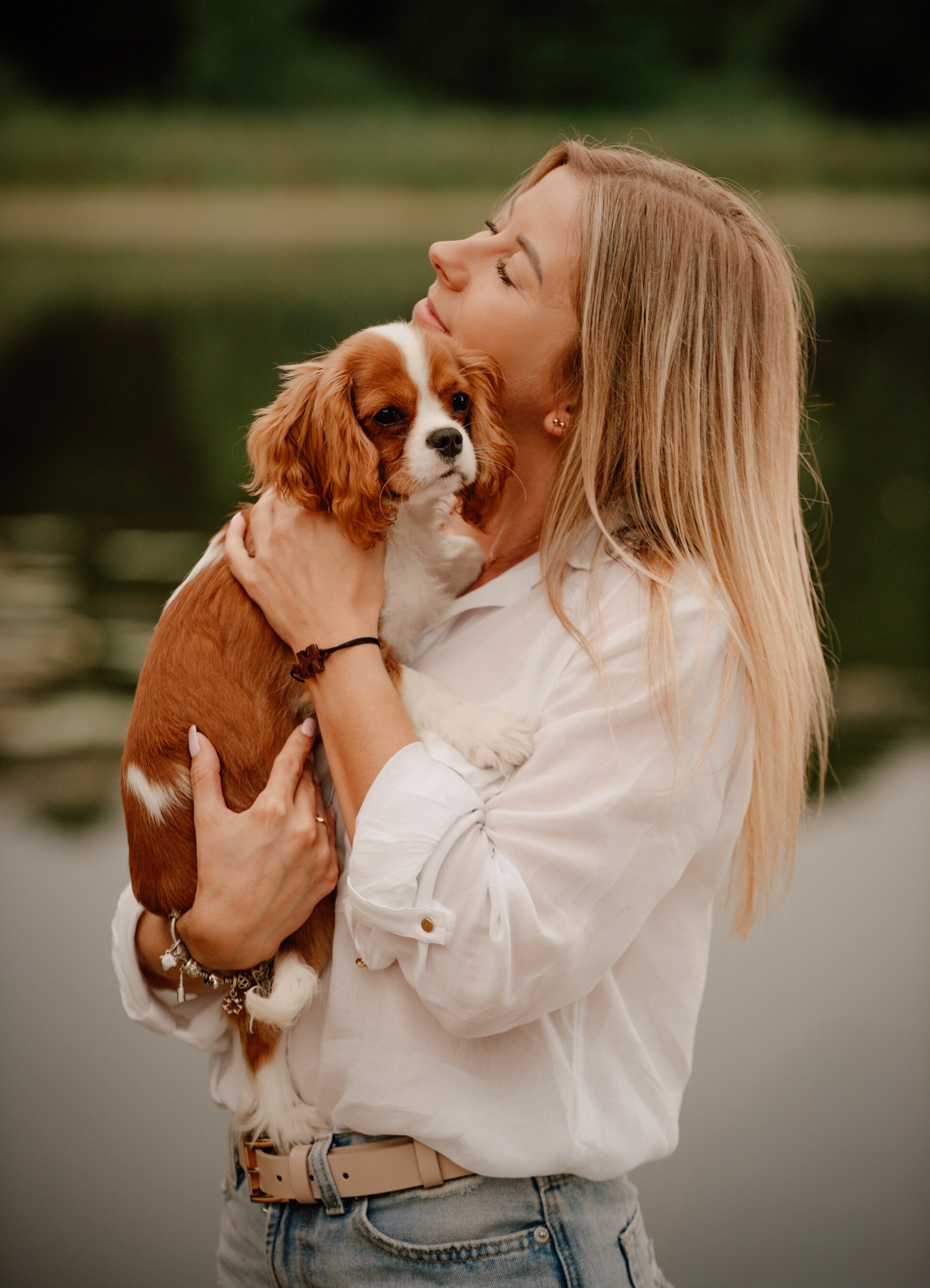Kobieta trzyma na rękach pieska cavaliera na tle jeziora podczas codziennej, luźnej sesji fotograficznej – fotografka Grudziądz