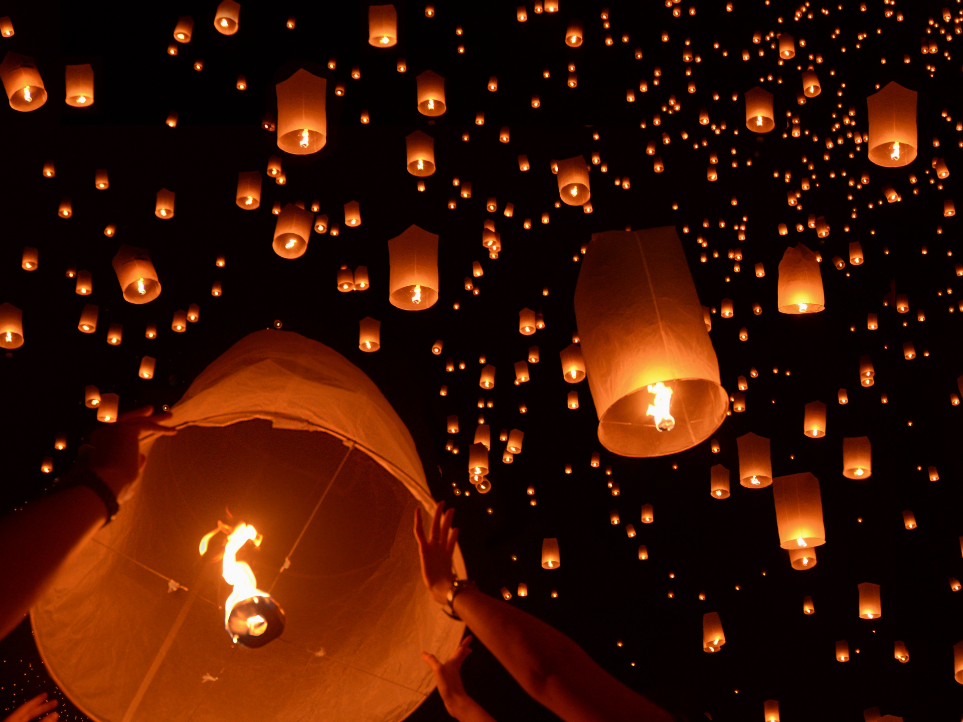 Honderden papieren lantaarns stijgen de nachtlucht in. Handen houden zo'n papieren lantaarn vast. Het is een schouwspel in Chiang Mai, Thailand. Het jaarlijks spektakel is Yi Peng. 