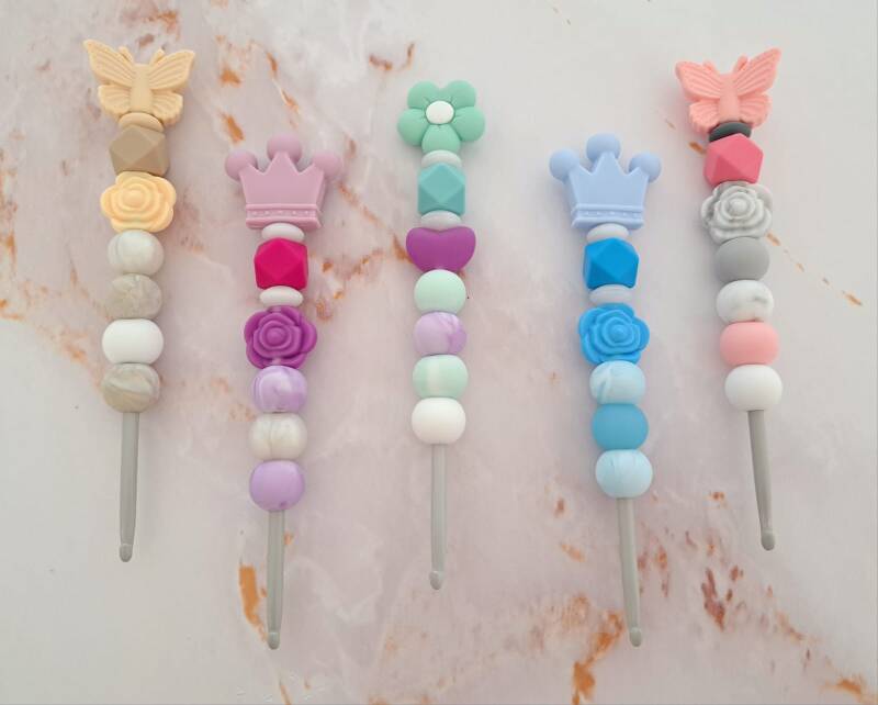 Das Bild zeigt fünf vertikal angeordnete Häkelnadeln der Größe 4,5mm mit individuell verzierten Griffen aus bunten Silikonperlen vor marmoriertem Hintergrund.