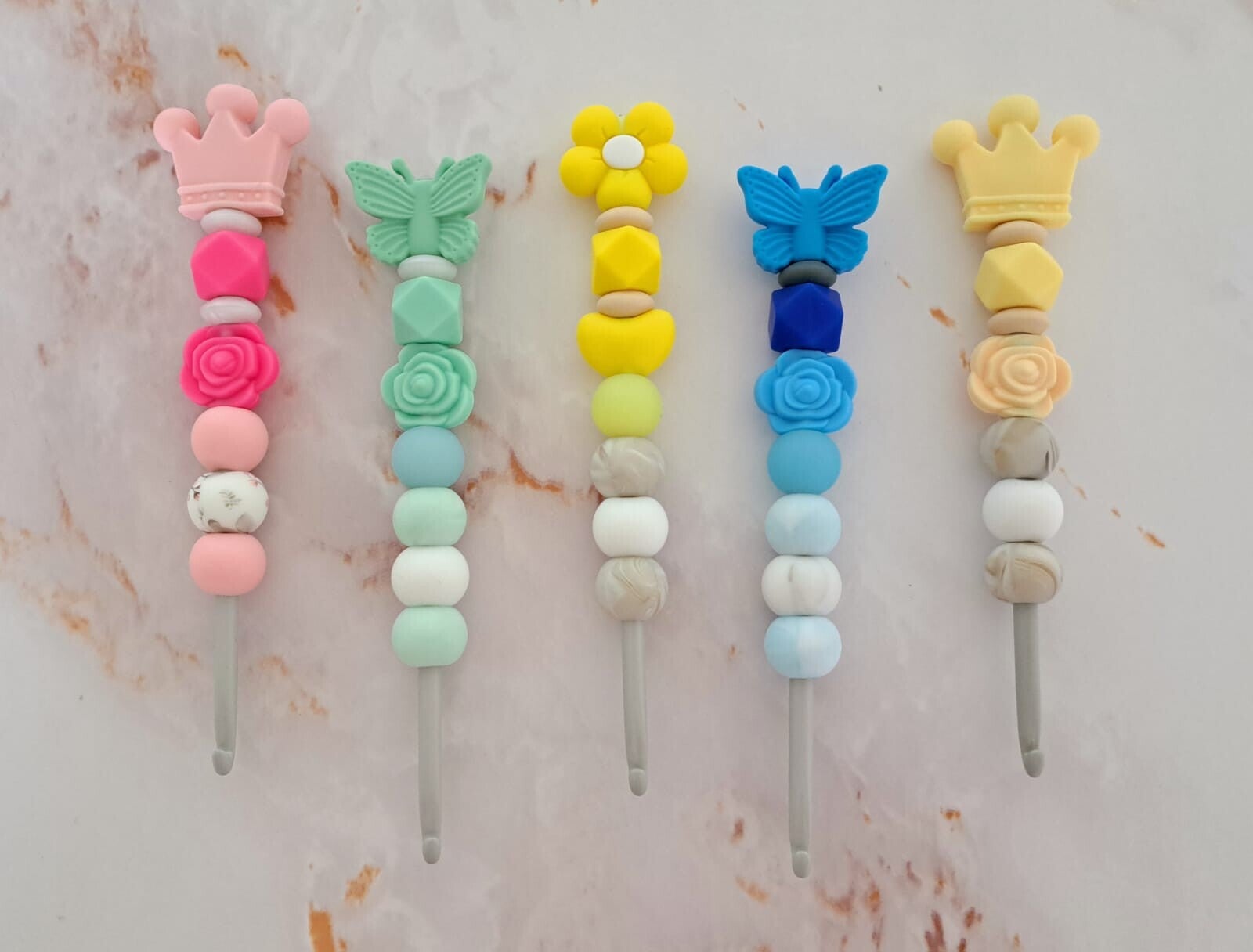 Das Bild zeigt fünf vertikal angeordnete Häkelnadeln der Größe 5,0mm mit individuell verzierten Griffen aus bunten Silikonperlen vor marmoriertem Hintergrund.