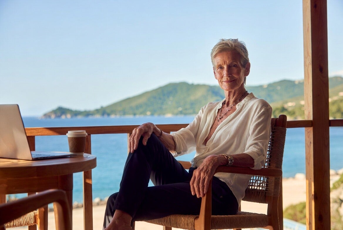 Confident woman in her 60s sitting at a sunny tropical resort with laptop, working on her online business
