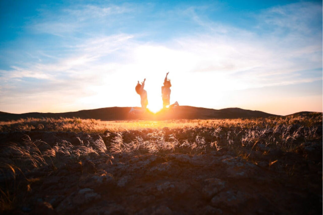 Foto von einer Wiese im Sonnenuntergang und zwei Freundinnen, die in größerer Entfernung vor der Sonne in die Luft springen.