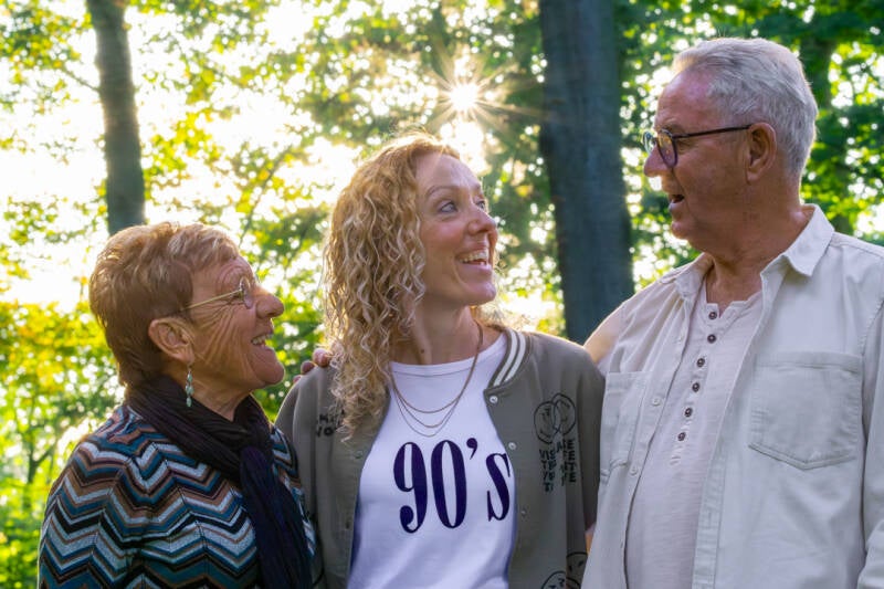 Liefdevol portret van een oudere vader en moeder met hun volwassen dochter, samen lachend in het bos.