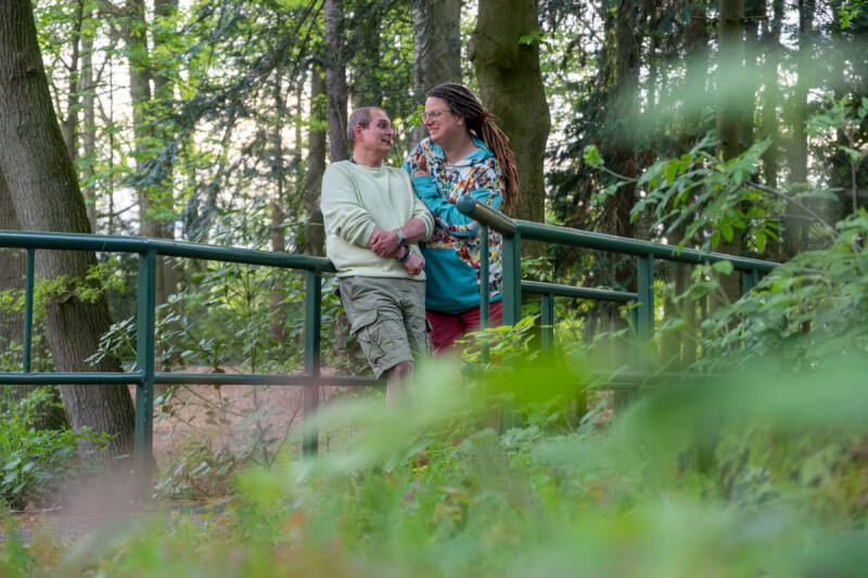 Koppel 50+ in beeld gebracht tijdens moment van verbinding op een bruggetje in een park.