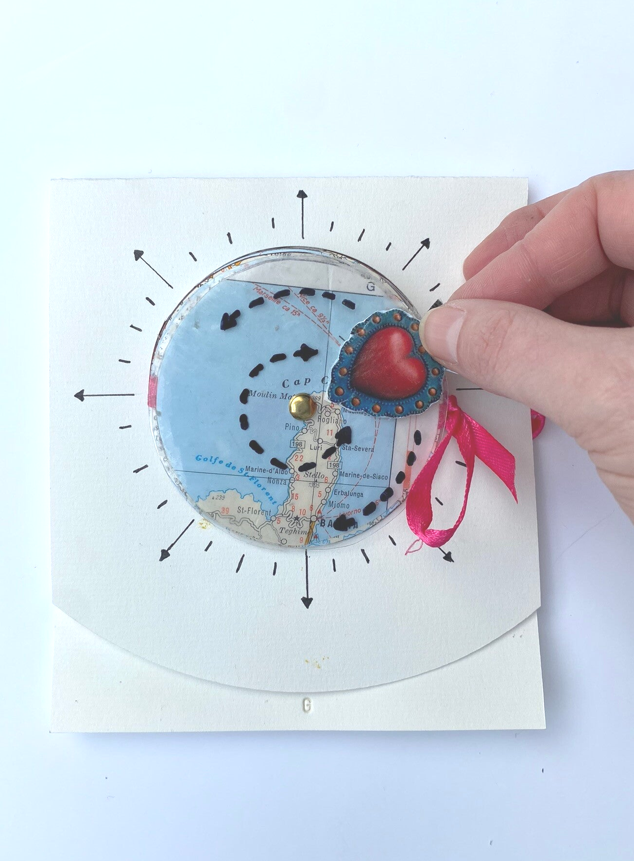 Op de foto zie je een handgemaakt kunstboekje met een ronde opening waardoor je een kompas ziet een aantal cirkels uit een oude landkaart geknipt die als een slinger aan een lint zijn bevestigd. Een hartje symboliseert de naald van de kompas. Als je de sl