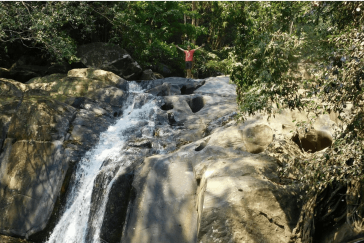 Pala-U Wasserfall in Thailand mit großen Felsen und herabfließendem Wasser, Mann steht oben am Wasserfall mit erhobenen Armen – Abenteuer und Naturerlebnis in tropischer Umgebung.