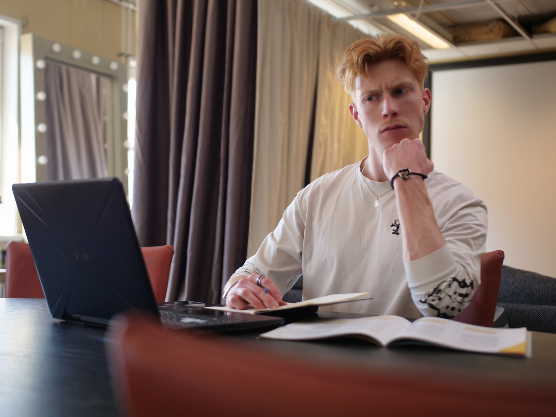 Roodharige man kijkt bedenkelijk terwijl hij aan een tafel zit met een laptop en opengevouwen boek.