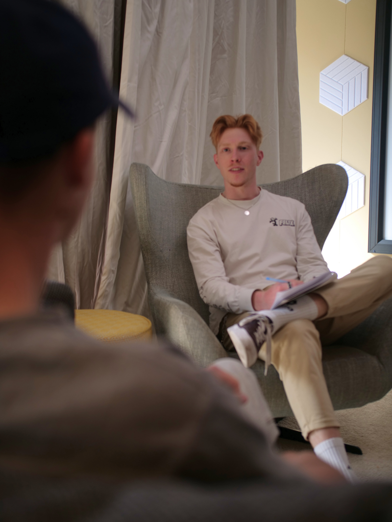 Een jonge man met rood haar zit in een grijze fauteuil en maakt aantekeningen tijdens een interview of gesprek.