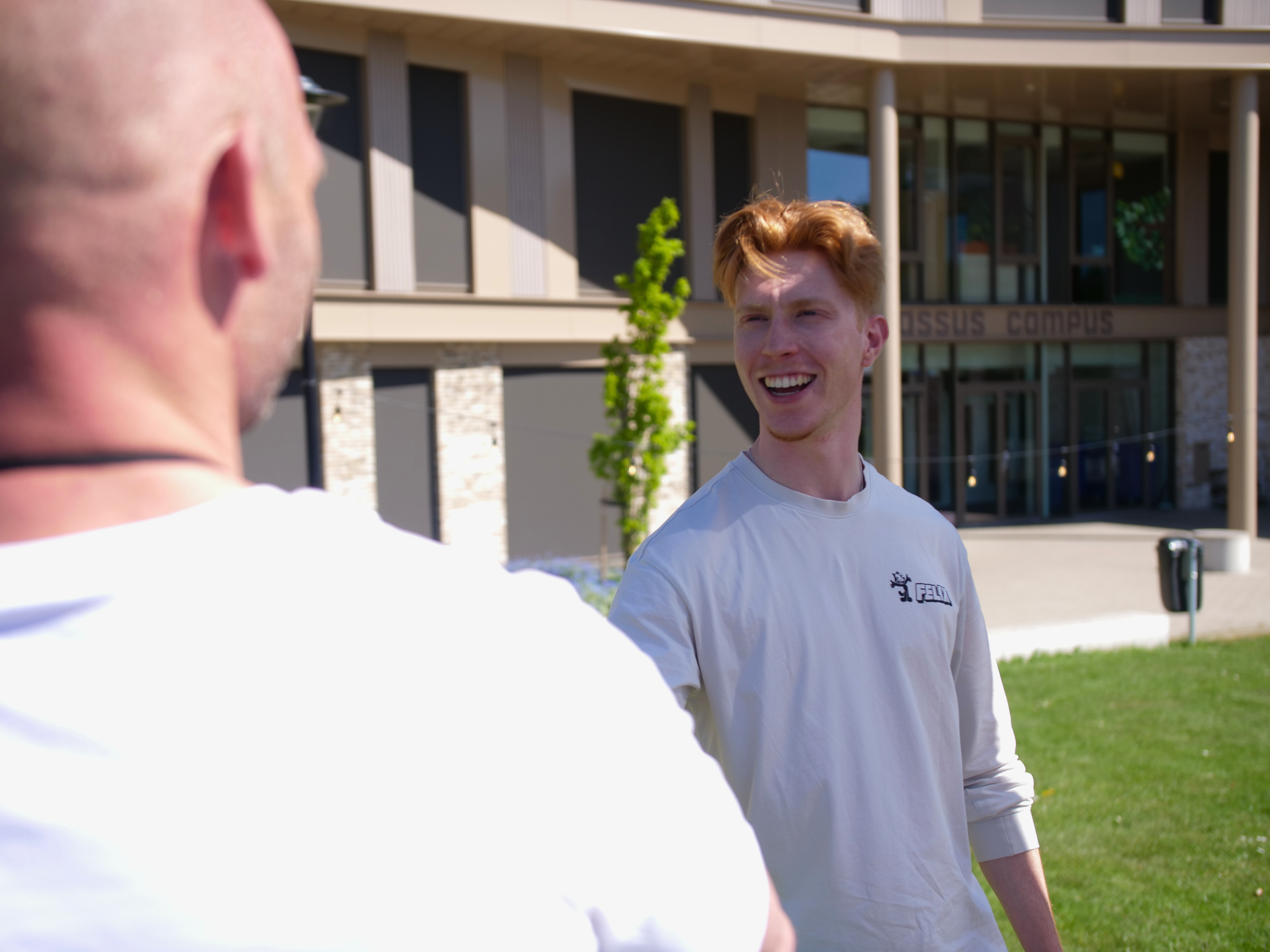 Lachende student schudt de hand van een andere persoon op een zonnige campus.
