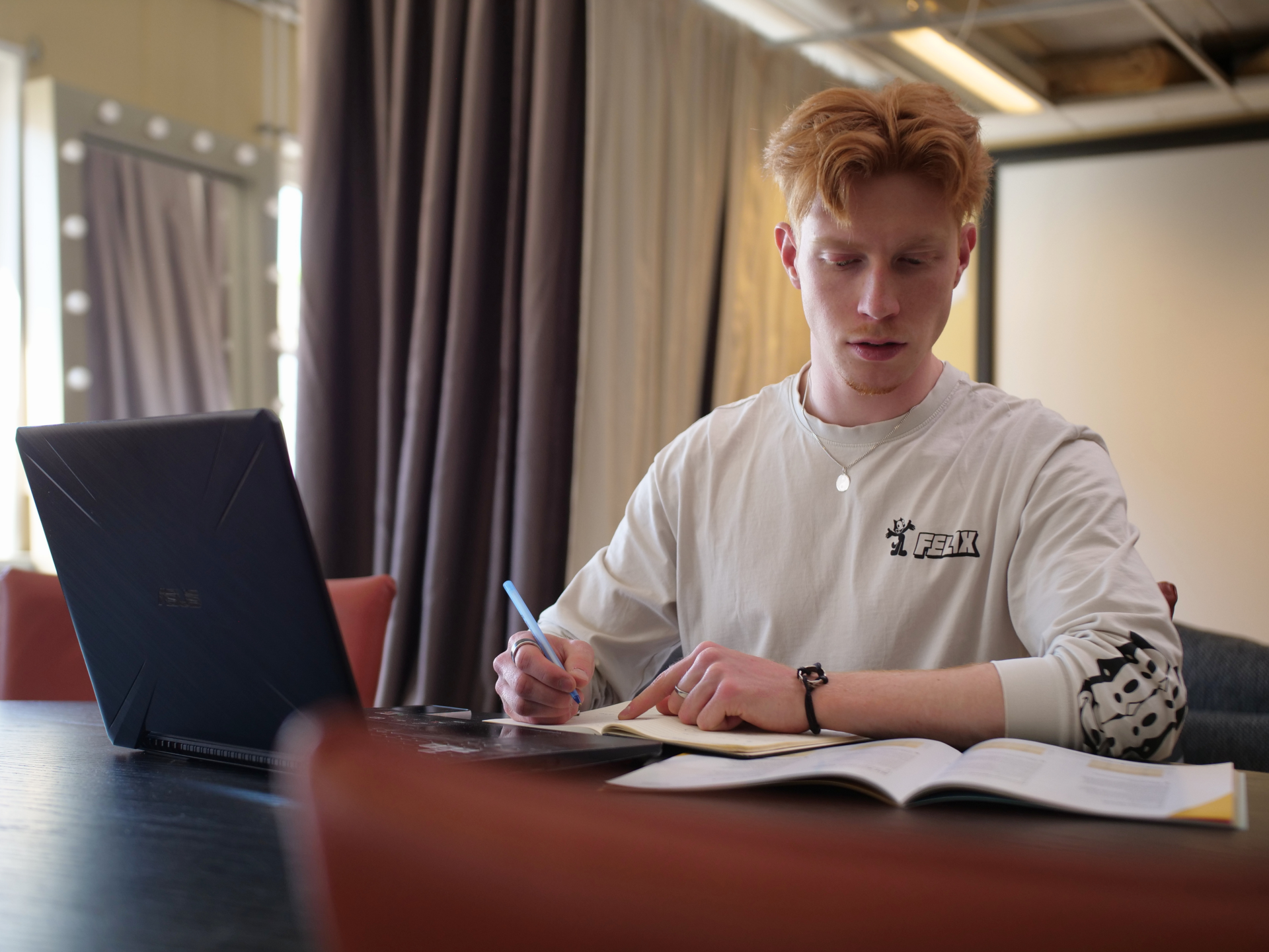 Roodharige man kijkt bedenkelijk terwijl hij aan een tafel zit met een laptop en opengevouwen boek.