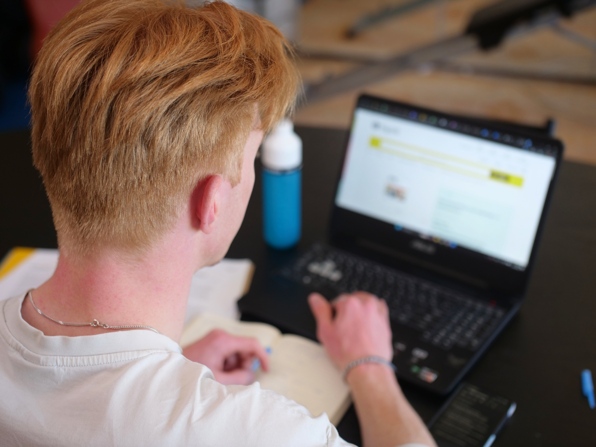 Achteraanzicht van een student die achter een laptop werkt met een blauwe drinkfles op tafel.