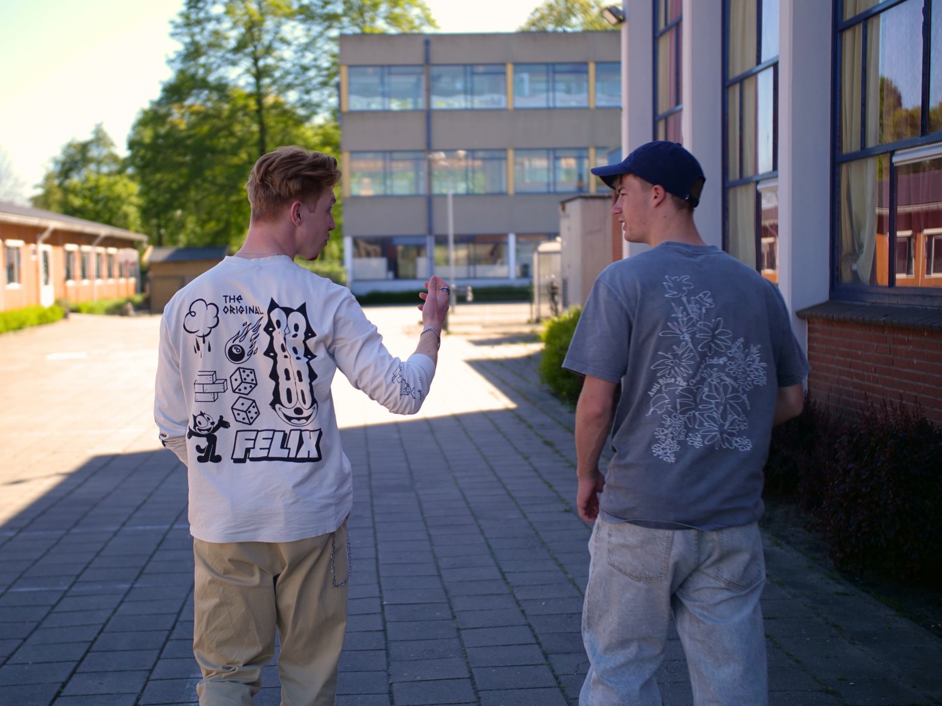 Een achteraanzicht van een leerling met zijn studiekeuzebegeleider die aan het praten zijn en die buiten aan het lopen zijn in de zon over een schoolplein