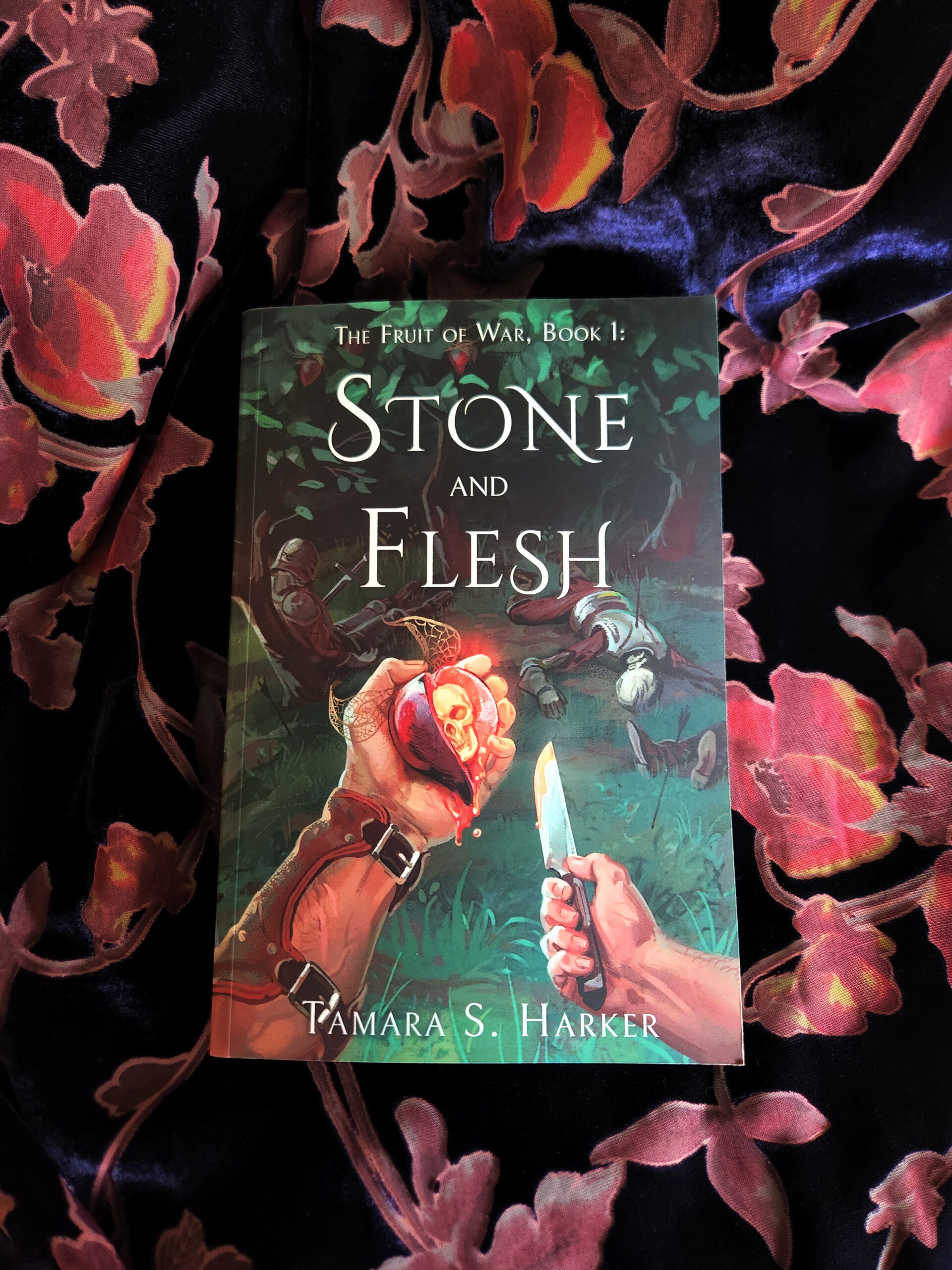 A book cover, titled "The Fruit of War, Book 1: Stone and Flesh". It is a dark green and black background with white text. A man's hand holds a purple fruit, cut open to reveal a skull inside. The book is laying on a purple and pink floral velvet fabric.