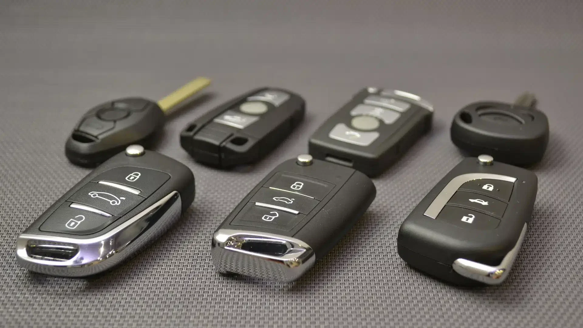 Mehrere moderne Autoschlüssel und Funkschlüssel verschiedener Automodelle, darunter Klappschlüssel, Keyless-Go Schlüssel und Fernbedienungen, nebeneinander auf einer strukturierten Oberfläche.