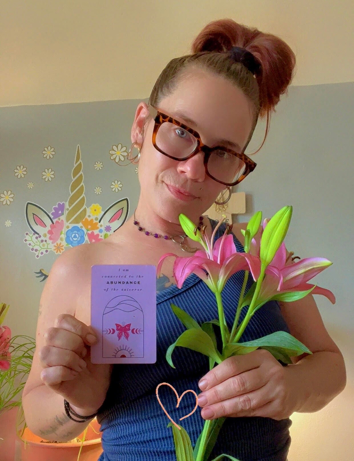 Crystal, The Illuminated Seeker, holding an affirmation card and pink lilies beside colorful unicorn artwork.