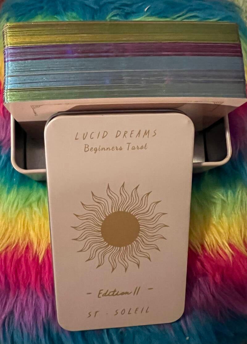 A close-up photo of the Atesswy Lucid Dreams Tarot Deck featuring vibrant rainbow-gilded edges, celestial-themed tarot cards, and a compact tin box for storage and protection.