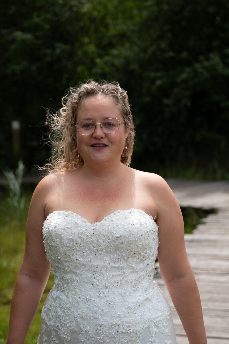Portret van een glimlachende vrouw in haar kanten trouwjurk aan het begin van een houten vlonderpad buiten in de natuur, gefotografeerd door Timeless Tales.