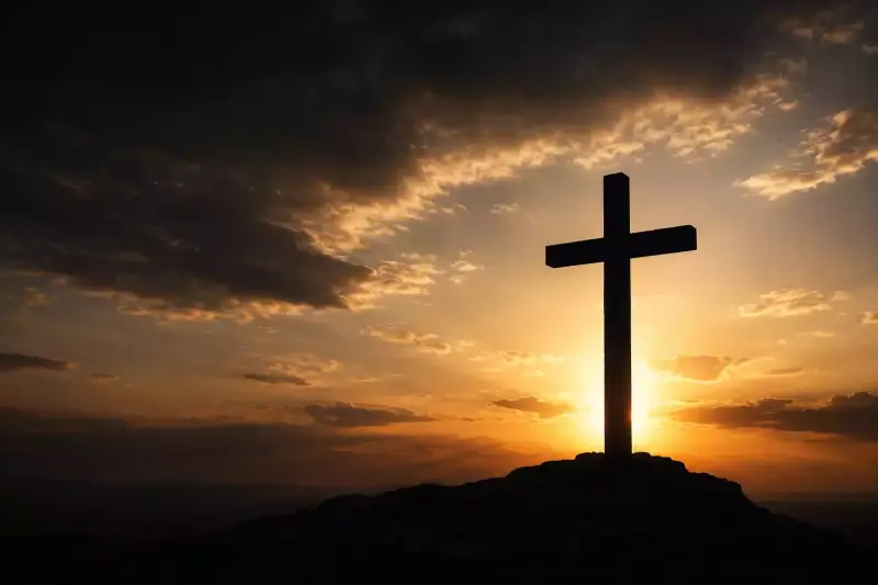 Silhouette d’une croix en contre-jour sur une colline, éclairée par une lumière traversant un ciel sombre en fin de journée