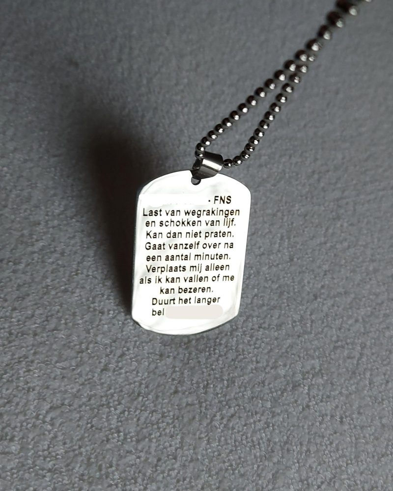 Medische SOS ketting achterkant. Tekst gegraveerd: Chloé Calis - FNS. Last van wegrakingen en schokken van lijf. Kan dan niet praten. Gaat vanzelf over na een aantal minuten. Verplaats mij alleen als ik kan vallen of me kan bezeren. Duurt het langer bel (