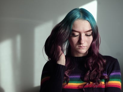 Person with teal and purple hair wearing a rainbow-striped sweater, looking down in soft lighting.