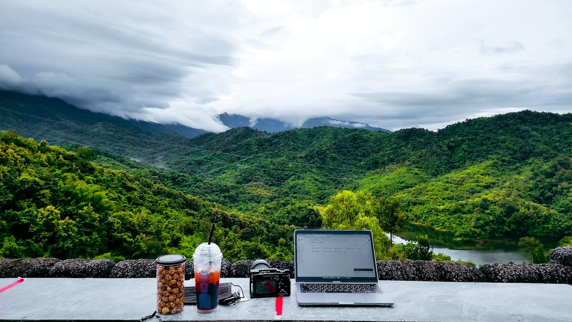 Station with PC, camera, drink in a glass and snacks, on a table that overlooks a beautiful panorama of a valley towards a river. Goal of every digital nomad.